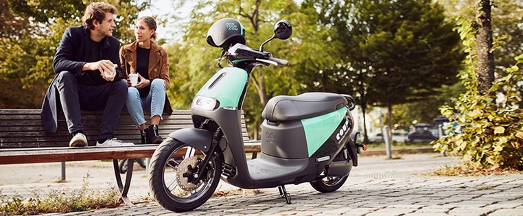 Mann und Frau sitzen auf Parkbank, vor ihnen steht ein E-Roller