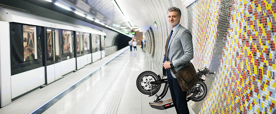 Mann steht an einer Ubahn-Haltestelle mit einem Elektrotretroller Metz Moover in der Hand