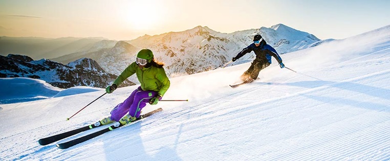 Zwei Skifahrer fahrer hintereinander auf einer leeren Piste im Gegenlicht