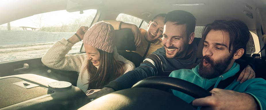 Gruppe junger Erwachsenen sitzen teilweise unangeschnallt im Auto und hören lachend Musik