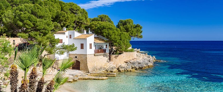 Reisen Spanien Mallorca, weißes Ferienhaus am Strans mit hellblauem Wasser