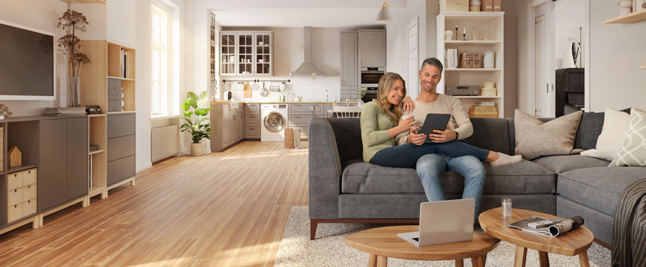 Junges Paar sitzt auf Sofa und schaut in ein Tablet, im Hintergrund Küche und Wohnzimmerschrank mit Hausrat